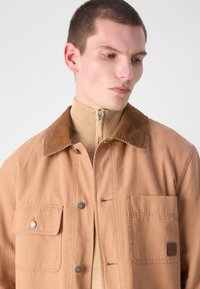 Lichtbruine jas met een corduroy kraag, twee knoopzakken op de borst en een gestructureerde gebreide coltrui. Bevat merkhardwaredetails.
