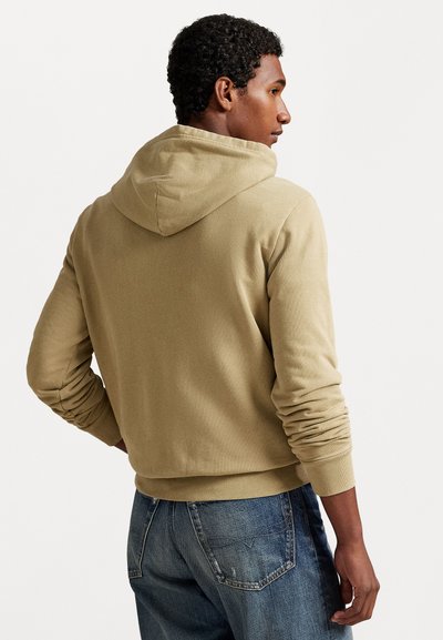Man met een beige hoodie en blauwe broek, zijwaarts gekeerd tegen een effen lichte achtergrond met handen op de heupen.