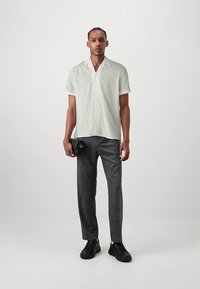 Homme portant une chemise blanche à manches courtes avec un motif en œillets, un pantalon gris, des baskets noires, tenant une pochette noire.