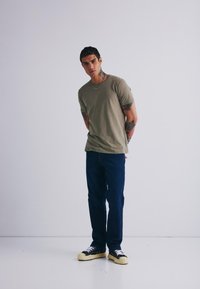Camiseta de cuello redondo verde oliva, jeans rectos azul, y zapatillas negras con suelas amarillas; tatuajes visibles en los brazos y el cuello.