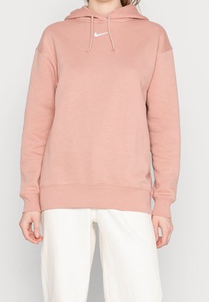 Personne portant un sweat à capuche rose Nike avec cordons et un pantalon blanc, debout devant un fond clair uni.