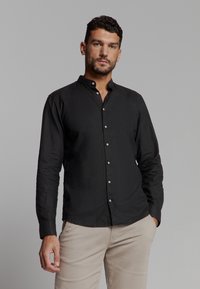 Zwarte langarmshirt met een mandarinakraag, voorzien van een knoopsluiting aan de voorkant en een gladde textuur. Gecombineerd met beige broek.