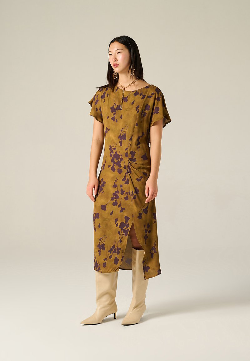 Braunes Blumenkleid in Midi-Länge mit kurzen Ärmeln, das einen seitlichen Schlitz und ein drapiertes Design aufweist. Kombiniert mit beigefarbenen Stiefeln mit Absatz bis zum Knie.