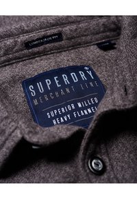 Camisa de franela en gris oscuro con un tejido texturizado, que presenta una etiqueta tejida en azul que dice "Superdry Merchant Line" y "Superior Milled Heavy Flannel."