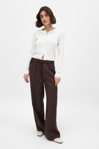 Weißer, langärmeliger Cardigan mit Knöpfen, kombiniert mit lockeren, braunen Hosen mit Kordelzug. Das Model trägt hellfarbene Schuhe und steht vor einem schlichten Hintergrund.