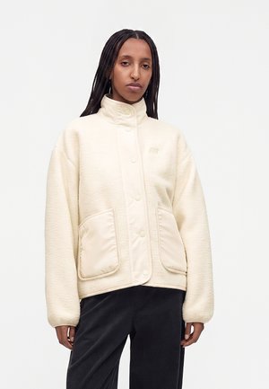 Femme aux cheveux tressés portant une veste polaire crème à col montant avec de grandes poches avant et un pantalon noir sur un fond uni.