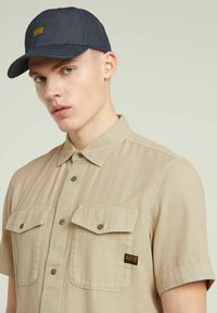 Beige overhemd met korte mouwen en een knoopsluiting aan de voorkant, voorzien van twee borstzakken, gecombineerd met een donkerdenim pet met een geel logo en blauwe stikdetails.