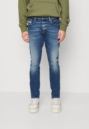 Mand iført slim-fit falmede blå jeans, hvide sneakers og en olivengrøn hættetrøje, stående mod en ensfarvet lys baggrund.