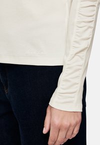 Cream long-sleeve shirt with gathered detailing on sleeves and embroidered logo near the hem, paired with dark denim jeans.