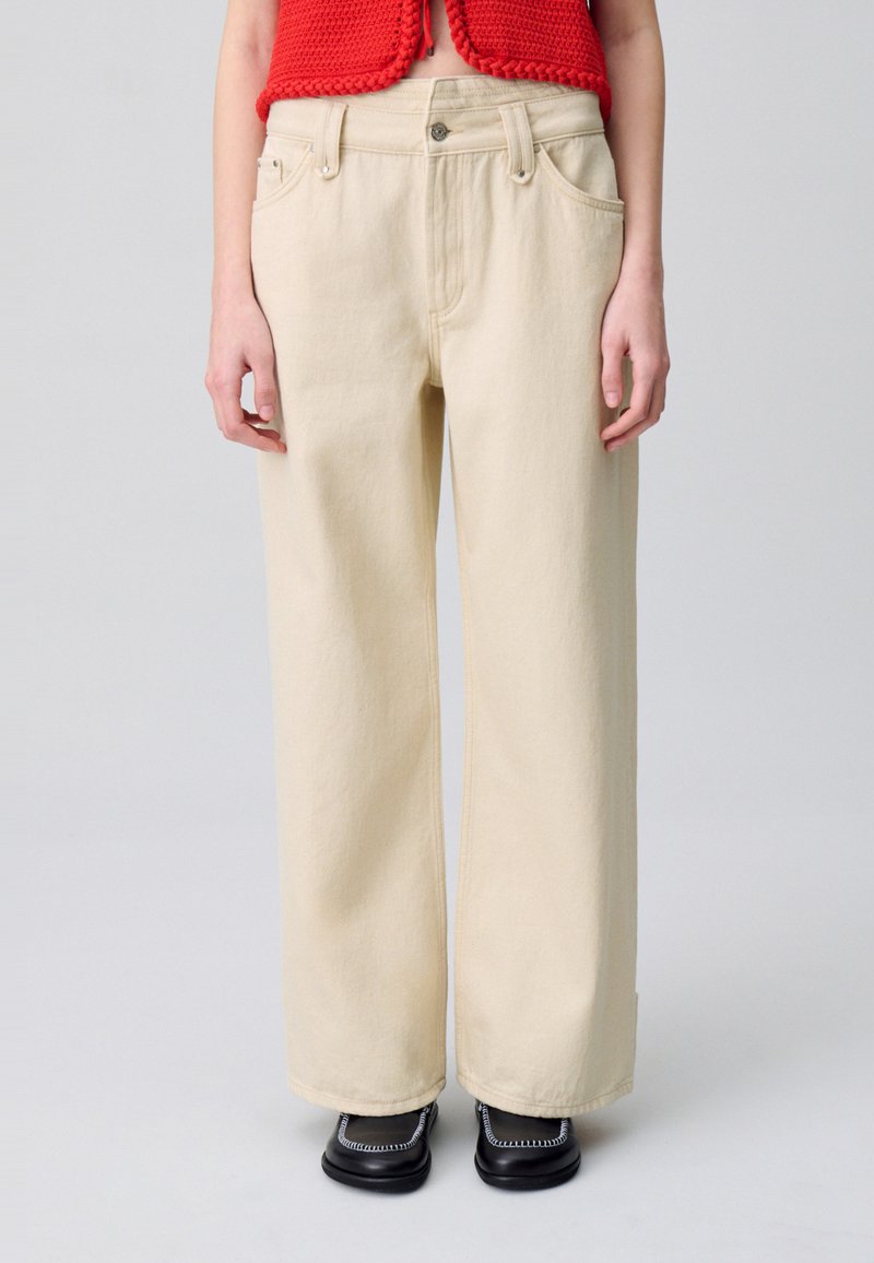 Pantalones de pierna ancha en algodón beige claro, con cintura alta, bolsillos frontales y una textura suave. Diseño minimalista con líneas limpias.
