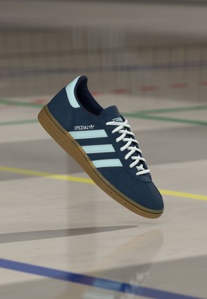 Baskets Adidas Spezial bleu marine avec bandes et lacets blancs, semelle marron, flottant au-dessus d'un terrain de sport intérieur avec des lignes peintes.