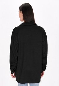Maglietta nera a maniche lunghe con un motivo geometrico testurizzato, colletto e polsini con bottoni. Taglio dritto, vista posteriore, indossata con jeans azzurri.