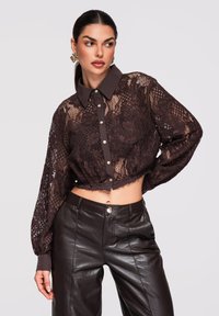 Femme portant un chemisier court en dentelle marron foncé avec des boutons et un pantalon en cuir taille haute, posant avec la main sur la hanche, fond clair.