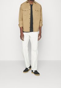 Hombre vestido con una camisa beige de botones sobre una camiseta negra, pantalones blancos y zapatillas negras con suela de goma, de pie contra un fondo claro.