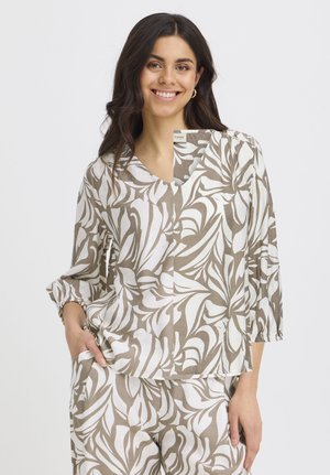 Usmievavá žena s dlhými tmavými vlasmi nosí zladenú blúzku béžovej a bielej farby s motívom listov, s trojštvrťovými rukávmi a voľné nohavice, pózujúc s rukou v vrecku.