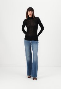 Svart langermet topp i et gjennomsiktig, teksturert stoff; parret med lyseblå vide jeans. Modellen står med hendene på hoftene mot en hvit bakgrunn.