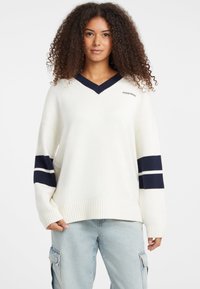 Fehér, oversized pulóver, navy V-nyakkal és csíkozott részletekkel az ujjakon. Anyaga kötött; bordás aljjal és hímzett logóval rendelkezik a mellrészen.