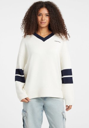 Maglione bianco oversize con scollatura a V blu navy e accenti a strisce sulle maniche. Il materiale è a maglia; presenta un orlo ribattuto e un logo ricamato sul petto.