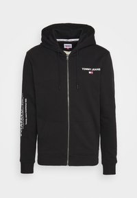 Haut zippé noir en mélange de coton, avec un logo sur la poitrine et du texte sur une manche. Il possède une capuche avec cordon de serrage et des poches avant.