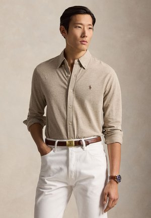 Mand iført beige langærmet polo med op rullede ærmer, hvide bukser, brun bælte og armbåndsur, stående med den ene hånd i lommen.