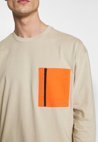 Beige hosszú ujjú ing, amelynek van egy téglalap alakú narancssárga zsebe fekete cipzárral. Puha tapintás és laza fazon.