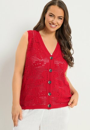 Lächelnde Frau mit langen Haaren, die eine ärmellose rote Häkelweste mit Knöpfen über einem weißen Oberteil und weißen Hosen trägt.