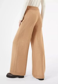 Hüftlange, camel-farbene Hose mit weitem Bein, geripptem Bund und strukturierten Seiteneinsätzen. Weicher Stoff und dicksohlige Schuhe vervollständigen den Look.
