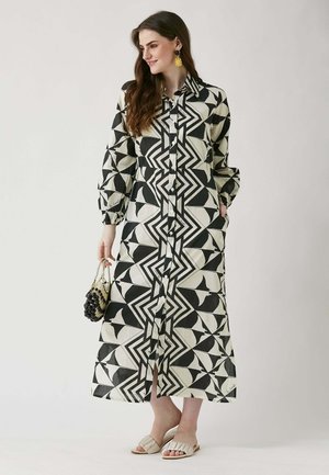 Bedrucktes, langes Hemdkleid mit schwarz-weißen geometrischen Mustern, Knopfleiste, langen Ärmeln und seitlichen Taschen. Tragen mit flachen Sandalen.