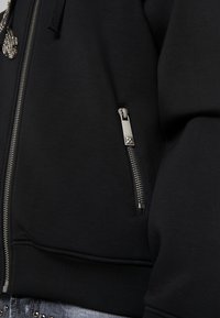 Sudadera negra con cremallera en la parte delantera que presenta un zipper metálico en el bolsillo izquierdo, tejido texturizado, y puños y dobladillo acanalados.