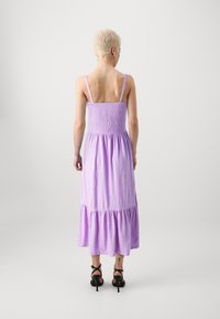 Vestido maxi sin mangas de color lila con un corsé fruncido, dobladillo en capas y textura de tela. Llevado con zapatos de tacón negros con tiras.