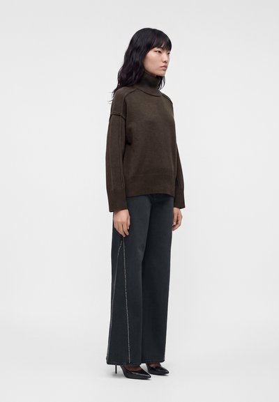 Femme aux cheveux noirs porte un pull à col roulé marron foncé, un pantalon large noir avec une couture latérale et des chaussures à talons hauts pointues noires.