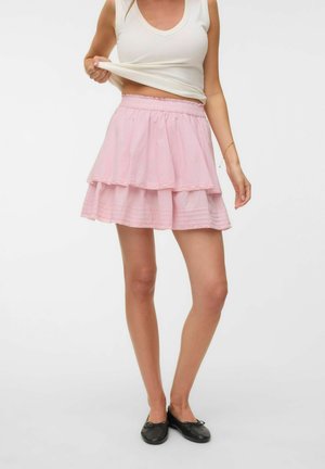 Femme portant un haut blanc sans manches et une mini-jupe rose à plusieurs couches, soulevant légèrement son haut, avec des ballerines noires, debout sur un fond blanc.