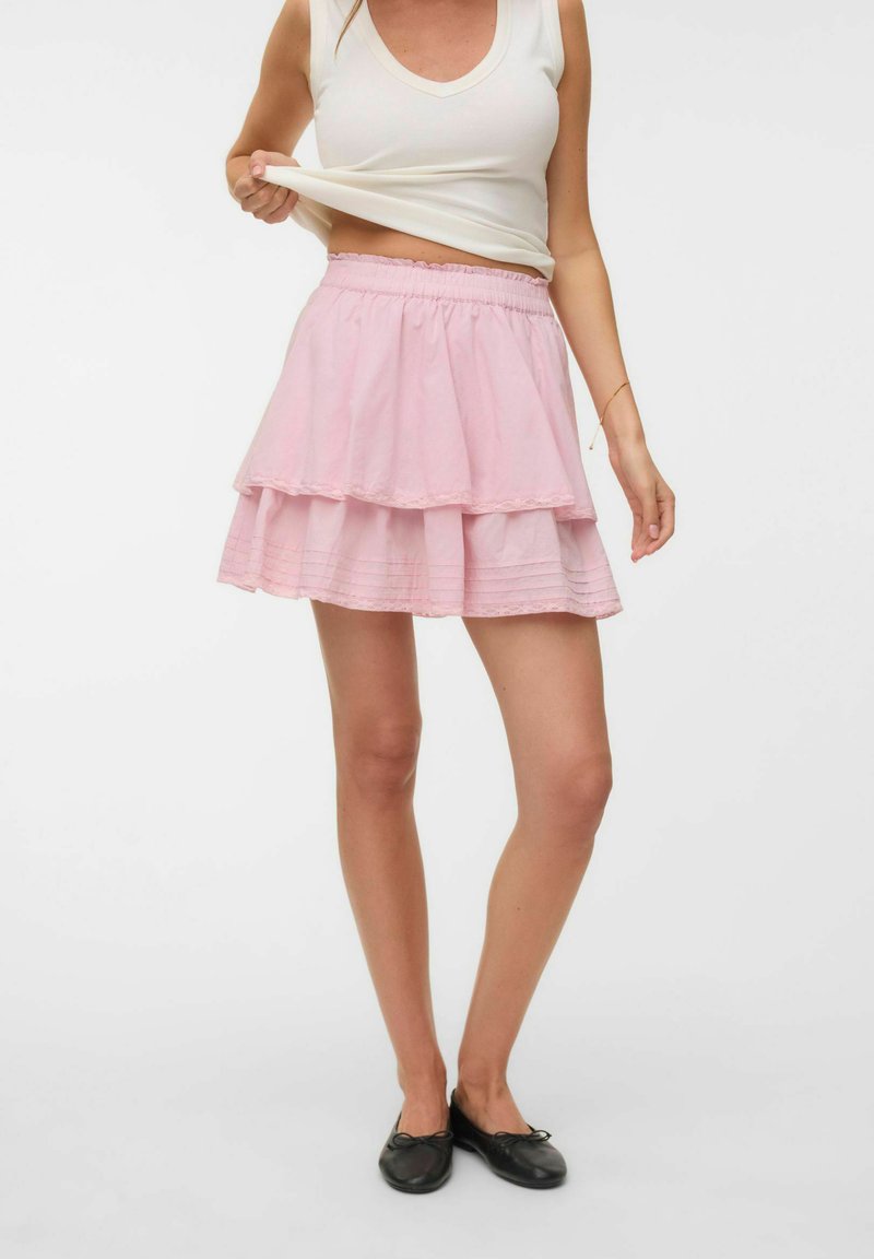 Femme portant un haut blanc sans manches et une mini-jupe rose à plusieurs couches, soulevant légèrement son haut, avec des ballerines noires, debout sur un fond blanc.