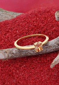 Goldring mit einem runden orangefarbenen Edelstein, der in vier Krallen gefasst ist, und auf einem rauen Zweig über rotem Granulat liegt.