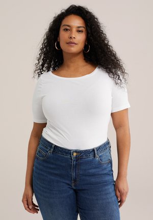 Vrouw met krullend haar, gouden oorringen, een witte top met korte mouwen en een blauwe spijkerbroek, staand tegen een effen achtergrond.