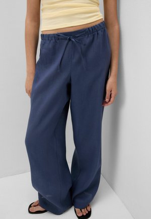 Osoba ubrana w luźne granatowe spodnie ze sznurkiem w talii, żółty krótki top i czarne japonki, stojąca przy białej ścianie.