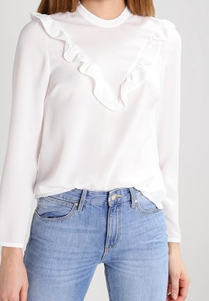 Femme portant un top blanc à manches longues avec un détail volanté sur la poitrine, associé à un jean taille haute bleu clair.