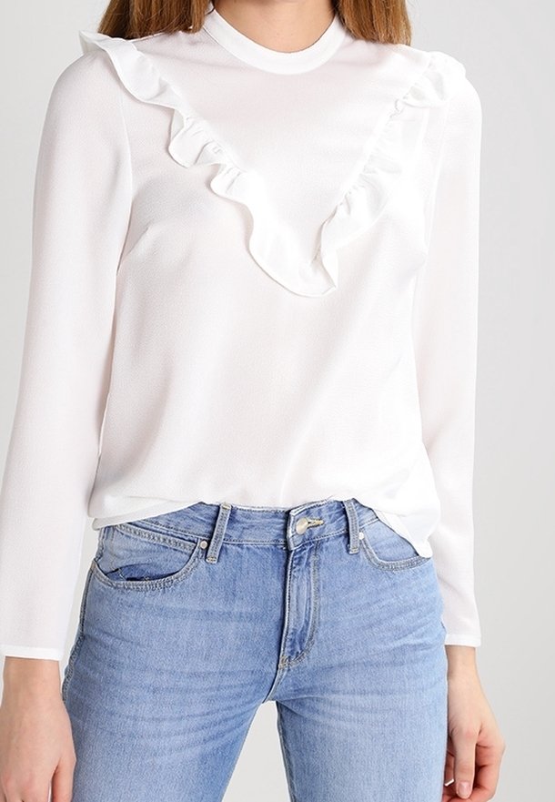 Femme portant un top blanc à manches longues avec un détail volanté sur la poitrine, associé à un jean taille haute bleu clair.