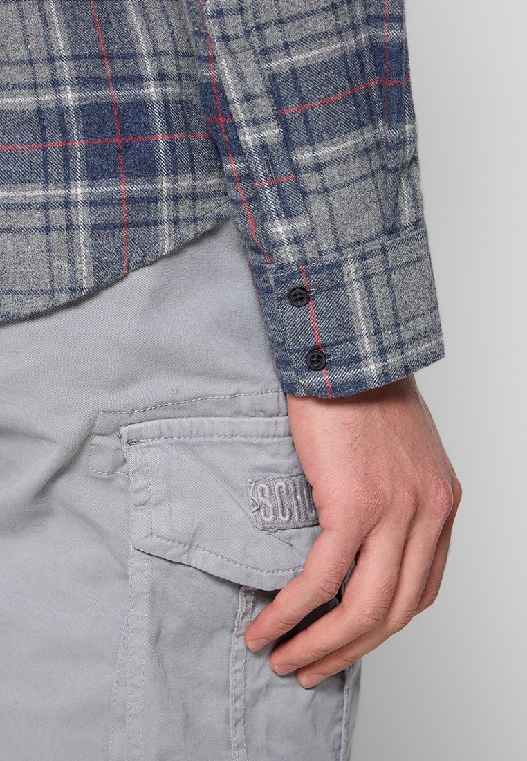 Szara koszula w kratę z niebieskimi i czerwonymi liniami, z zapinanym mankietem. W zestawie z jasnoszarymi spodniami cargo z kieszenią po bokach.
