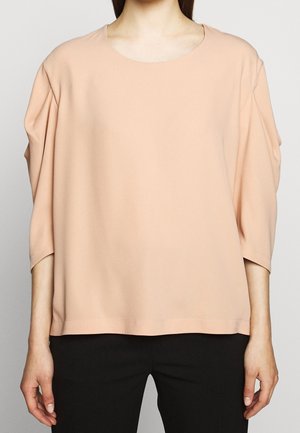 Woman wearing a loose, peach-colored long-sleeve top with dropped shoulders and black pants on a light gray background.