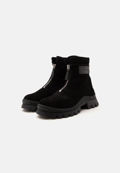Paire de bottines en daim noir avec semelles en caoutchouc épaisses et fermetures éclair argentées à l'avant sur fond blanc.