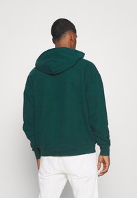 Felpa verde con cappuccio ampio, polsini e orlo a costine. Tessuto morbido; vestibilità rilassata con dettagli di cucitura visibili nelle cuciture.
