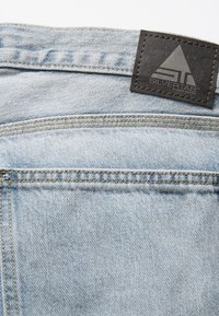 Világoskék farmeranyag, amely texturált felülettel rendelkezik, bőrből készült "SILVERTAB" felirattal ellátott folttal és ellentétes színű varrással a szélek mentén.