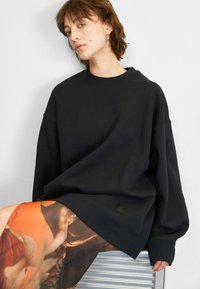 Sudadera negra de corte oversize en un material suave con hombros caídos, que presenta un pequeño parche de logo. Combinada con una falda transparente y estampada.