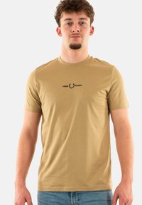 Camiseta de algodón color tostado con mangas cortas, escote redondo y un logo negro de "FRED PERRY" sobre una corona de laurel en el pecho.