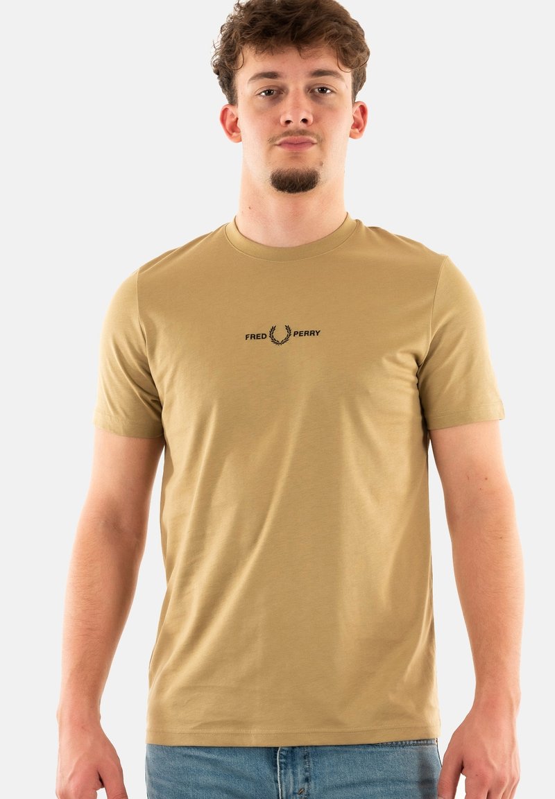 Camiseta de algodón color tostado con mangas cortas, escote redondo y un logo negro de "FRED PERRY" sobre una corona de laurel en el pecho.