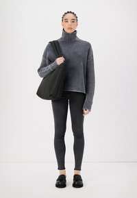 Siv pulover s rebrasto strukturo in visokim ovratnikom, črna obložena torba, temno ozke kavbojke in črne čevlje. Model stoji ob ozadju v beli barvi.