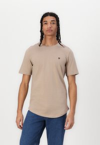 Beige trumpas rankovėmis marškinėliai, pagaminti iš medvilnės, su apvalia iškirpte. Ant kairiojo krūtinės kampo yra mažas juodas logotipas, o apatinė dalis tiesi.