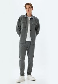 Grijze shirtjacket met twee borstzakken, gedragen over een witte t-shirt en grijze taps toelopende broek. Gecombineerd met witte sneakers. Glad oppervlak.