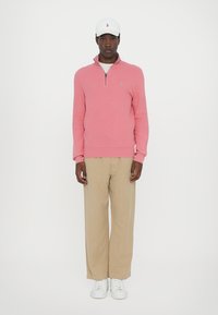 Sweat-shirt zippé rose clair et pantalon large beige. Casquette blanche avec logo et baskets blanches. Textures de tissu lisses et design simple.